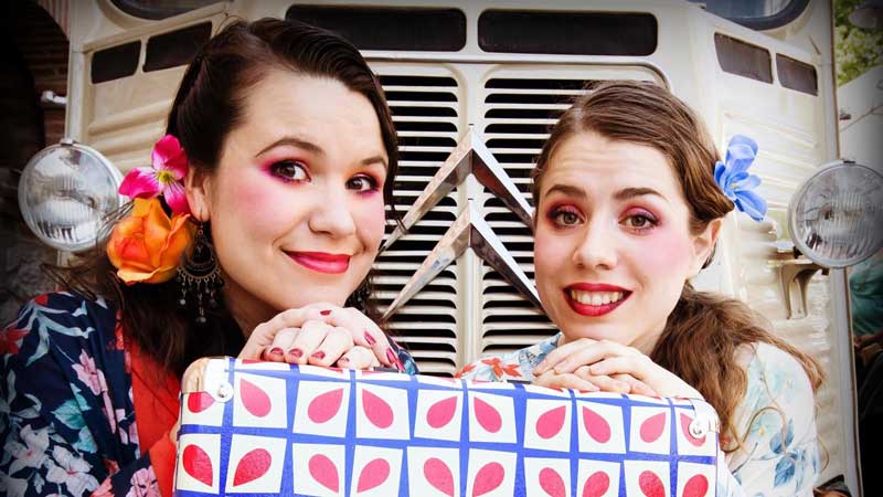 Las dos actrices con la cabeza apoyada en una maleta de colores