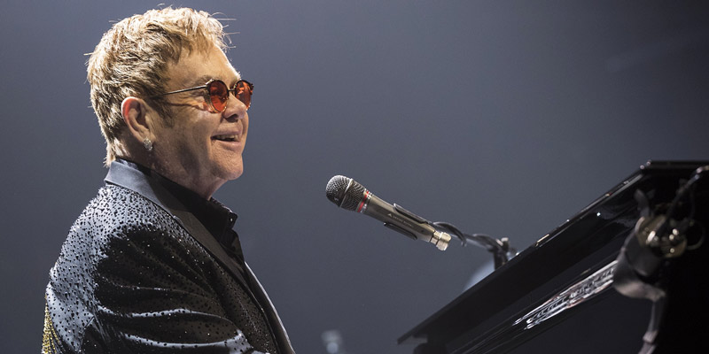 Elton John con gafas rojas con un micrófono y tocando el piano