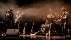 Concierto de músicos con una cantante, dos guitarristas y un bateria sobre un fondo negro