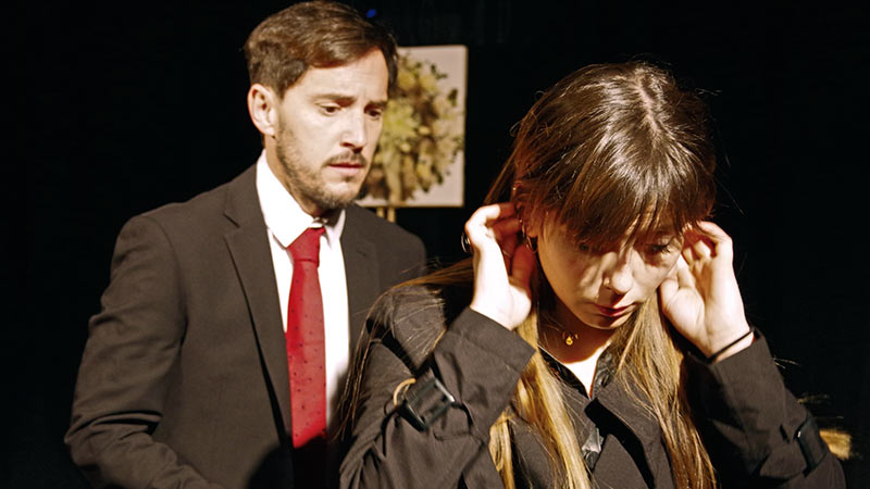 La pareja de actores en Incondicionales, hablan entre sí, ella le da la espalda a él mientras se recoge el pelo detrás de la oreja. Él tiene cara de arrepentimiento