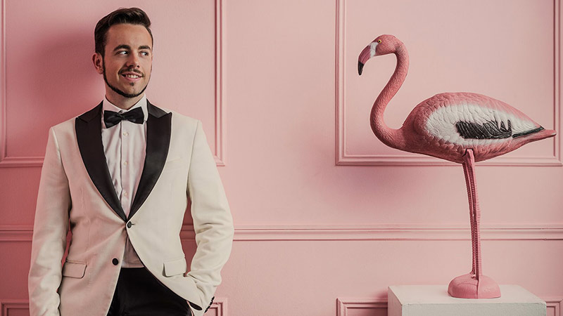 El Mago Marín posando junto a un flamenco de plástico en una habitaición rosa, lleva puesto un traje blanco 