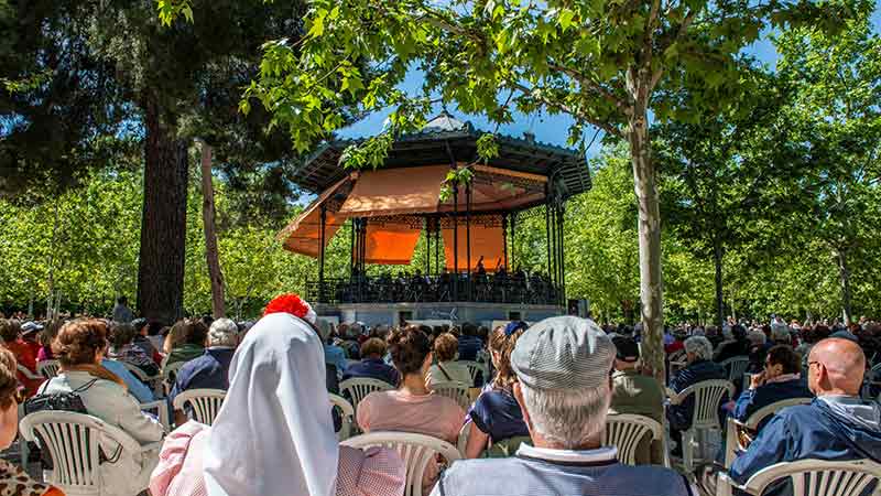 Concierto de la orquesta sinfónica en el parque de las vistillas, una pareja vestida de chulapos y chulapas en el centro, todos los asientos están llenos